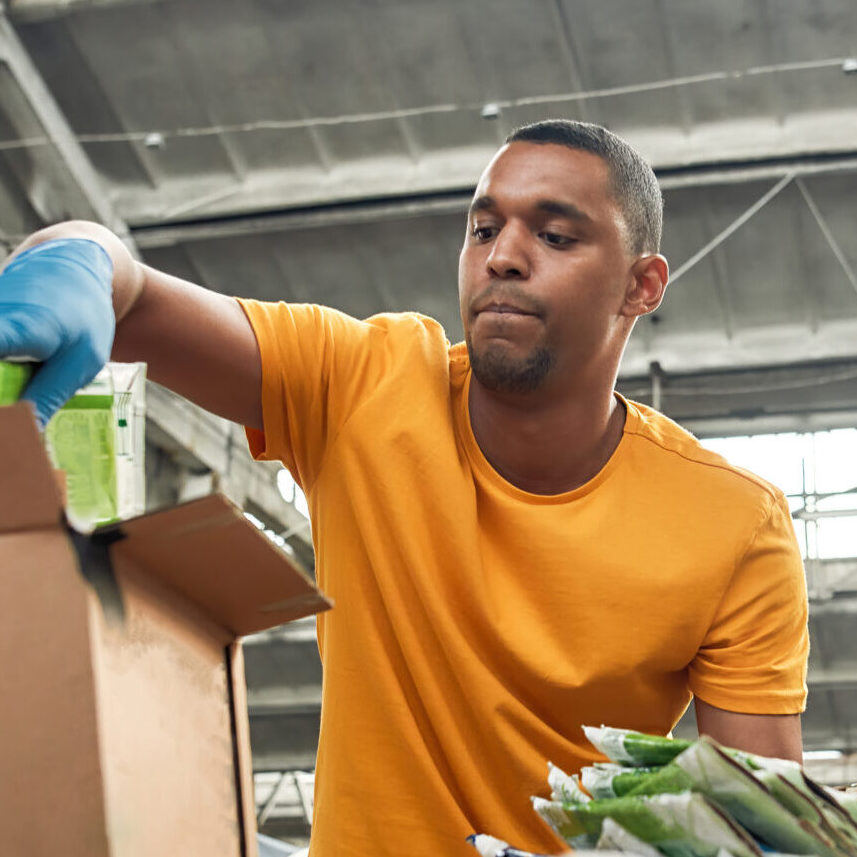 Young african american man packing boxes with waste paper for reuse while working on rubbish station, widescreen. Garbage sorting and recycling concept